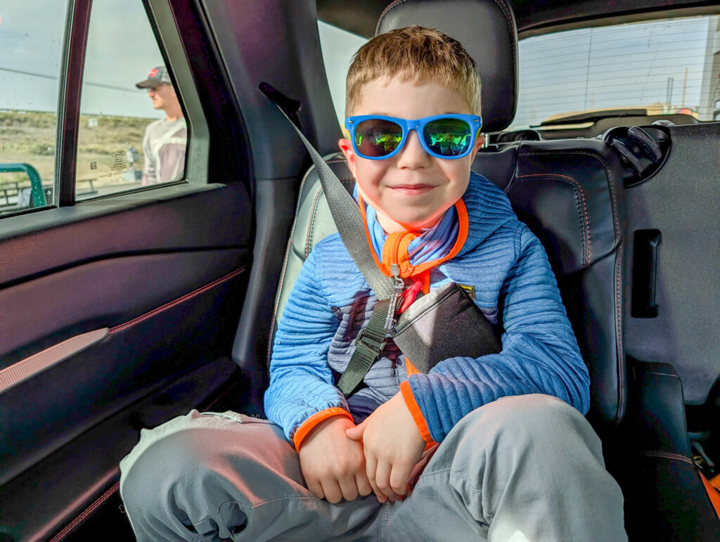 Young boy wearing blue sunglasses and striped shirt sits buckled in car seat during daytime road trip