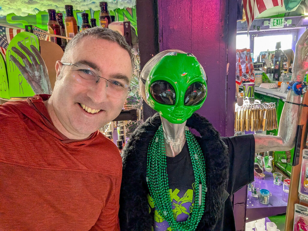 A man in a red shirt poses with a green alien mannequin inside a heavily themed extraterrestrial novelty shop in Moab, Utah.