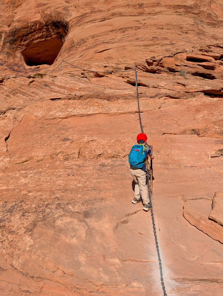 A hiker in a red helmet and blue backpack climbs a near-vertical sandstone slab using a fixed chain handrail in Fiery Furnace, Arches National Park.