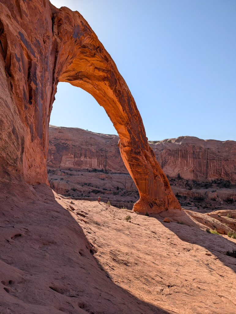 A hiker stands at the base of Corona Arch near Moab, dwarfed by the sweeping red sandstone span overhead.
