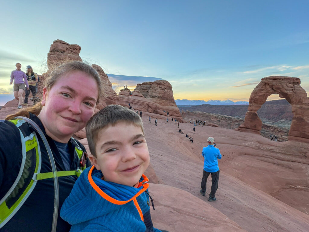 A woman and young boy take a selfie near Delicate Arch at golden hour, with the iconic sandstone arch and snow-capped La Sal Mountains behind them.