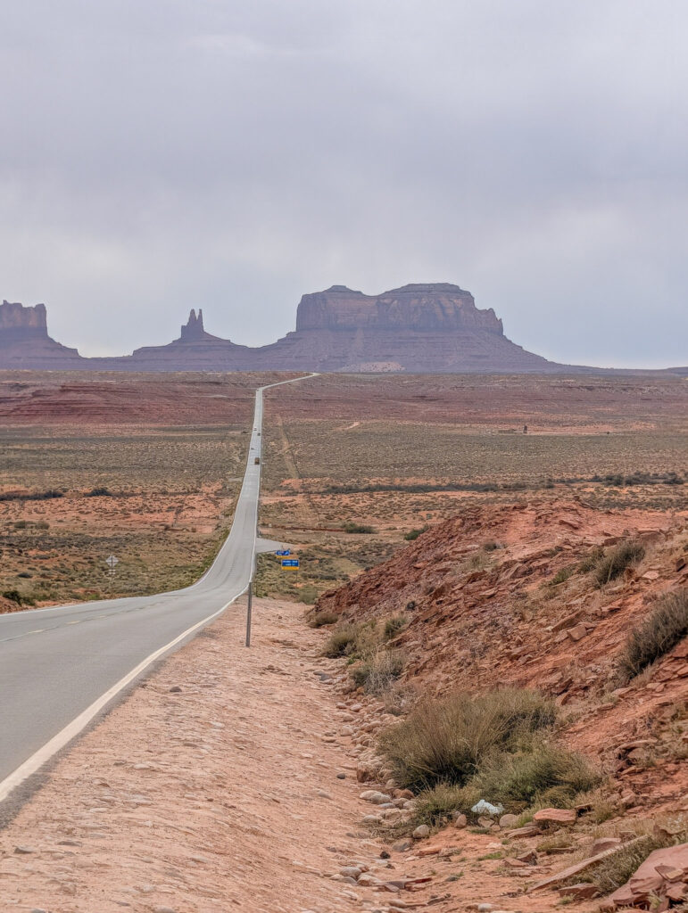 US Highway 163 stretches toward Monument Valley's sandstone buttes under an overcast sky, with red embankments lining the roadside.