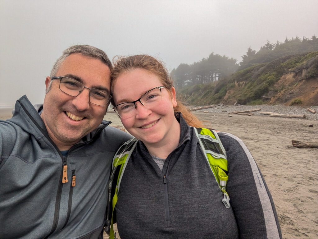 Two hikers taking a selfie on a foggy Pacific Northwest beach with forested bluffs and driftwood in overcast light