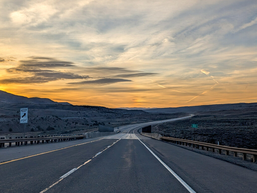 Highway crossing the Price River at golden hour, with rolling high-desert hills and a sunrise sky along US-6 in central Utah.