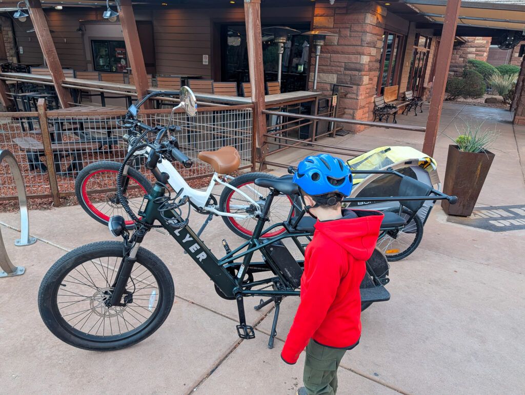 Child in a blue helmet stands beside a black Flyer electric cargo bike with a Burley trailer attached at a Springdale, Utah rental shop.