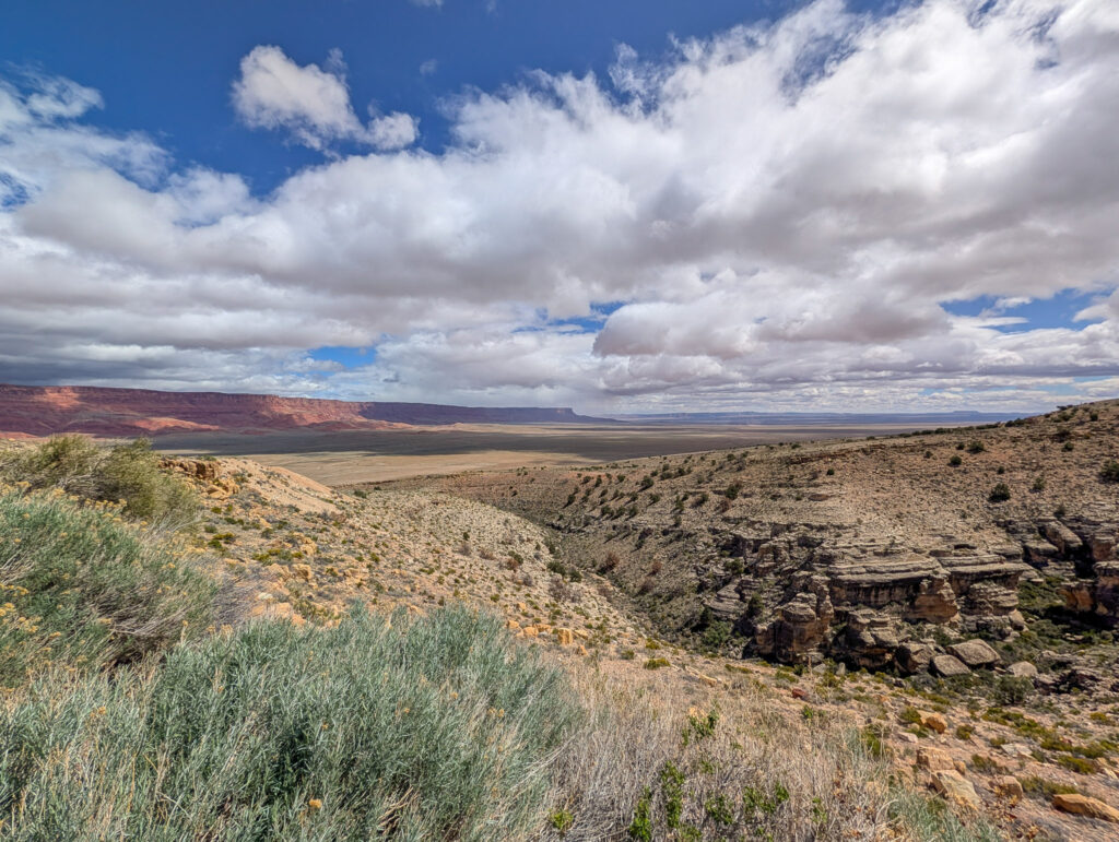 View from House Rock Valley Overlook across a wide arid valley toward the red-orange Vermilion Cliffs escarpment under dramatic cumulus clouds.