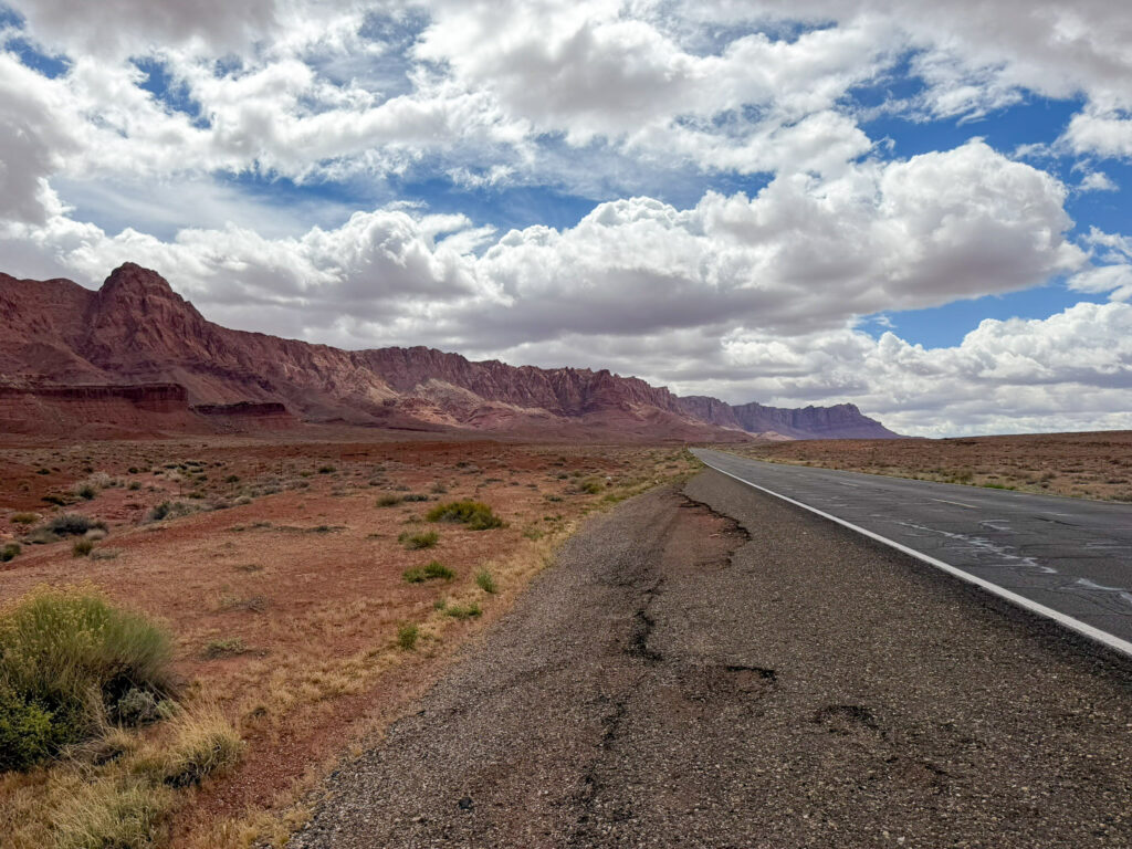 US-89A stretches toward the horizon beneath cloudy skies, flanked by the red sandstone Vermilion Cliffs and desert scrub in northern Arizona.