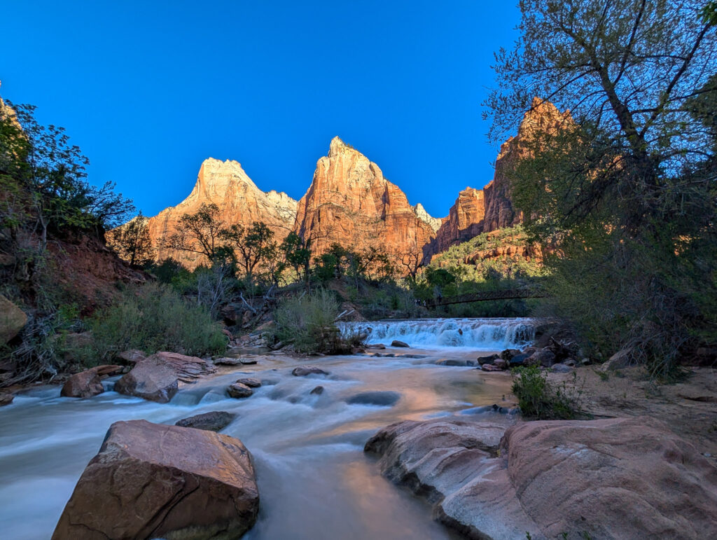 Long-exposure shot of the Virgin River cascading over rocks below the Court of the Patriarchs' three sunlit sandstone peaks in Zion National Park.