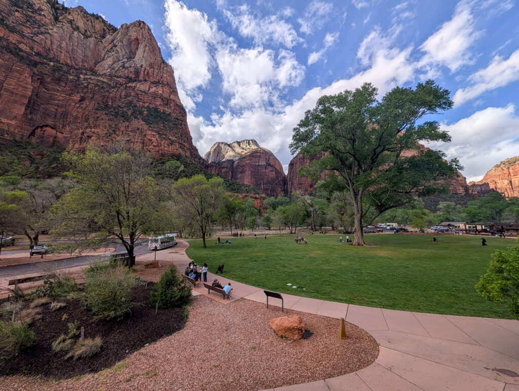 Visitors relax on the Zion Canyon Visitor Center patio lawn beneath towering red sandstone cliffs at golden hour.