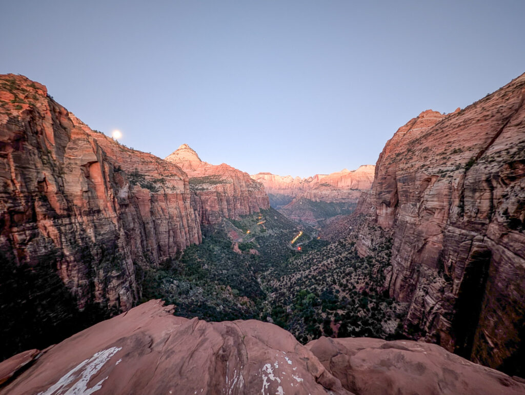 A nearly full moon sets over Zion Canyon at dawn, seen from Canyon Overlook Trail, with alpenglow on the Navajo sandstone walls below.