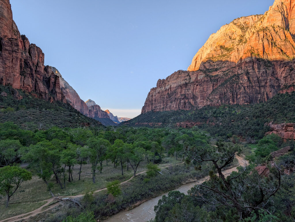 Zion Canyon valley view with the Virgin River winding past cottonwoods beneath warm alpenglow on the Navajo sandstone walls.