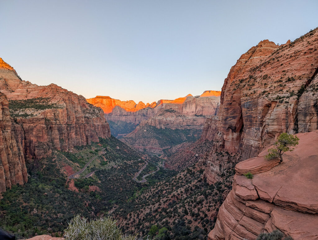 Warm sunrise light bathes the Towers of the Virgin and Zion Canyon as seen from Canyon Overlook Trail, with the winding Zion–Mount Carmel Highway visible far below.
