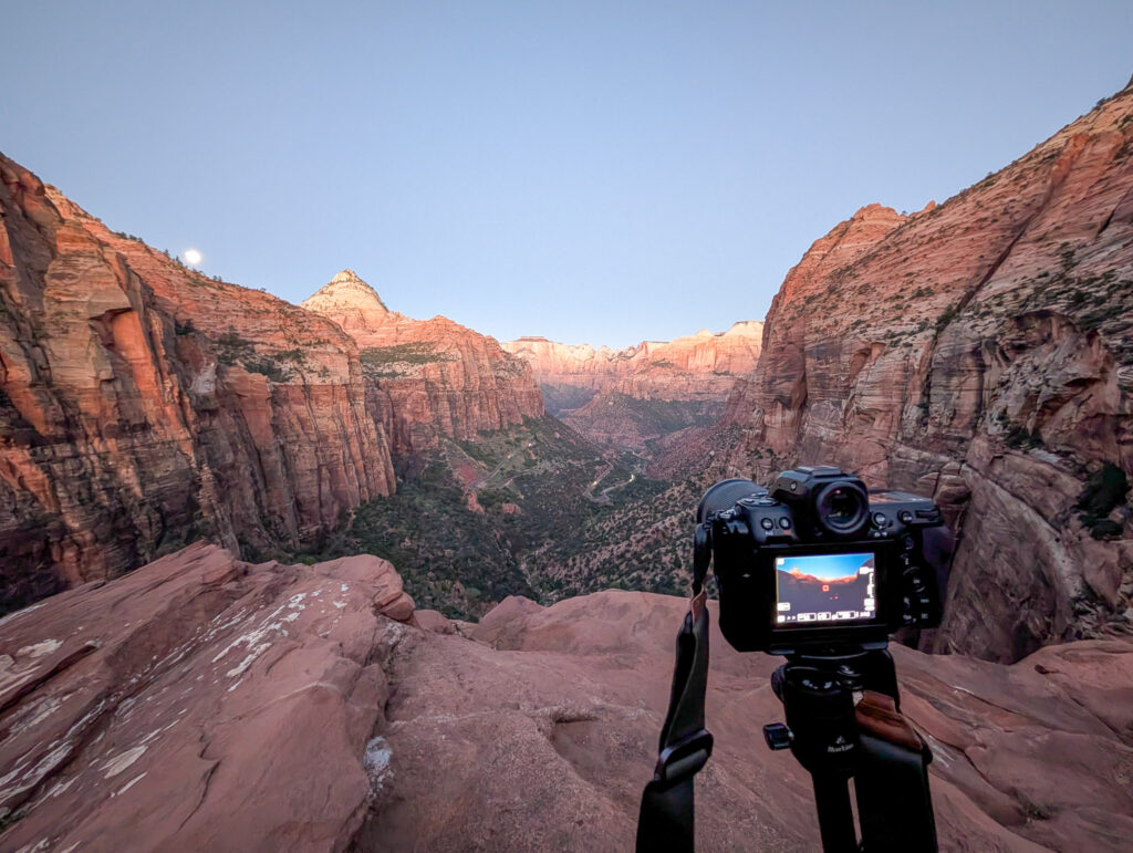 Camera on a tripod at Canyon Overlook captures Zion Canyon at dawn, with alpenglow on sandstone walls and a full moon above the rim.
