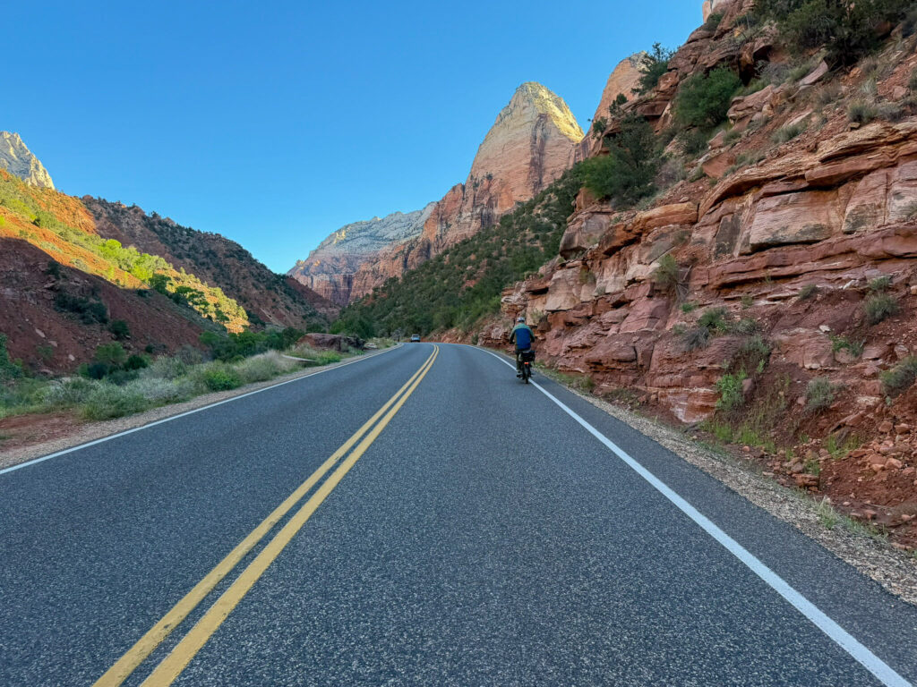 A loaded touring cyclist rides along Zion Canyon Scenic Drive beneath towering red Navajo sandstone cliffs under a clear blue sky.