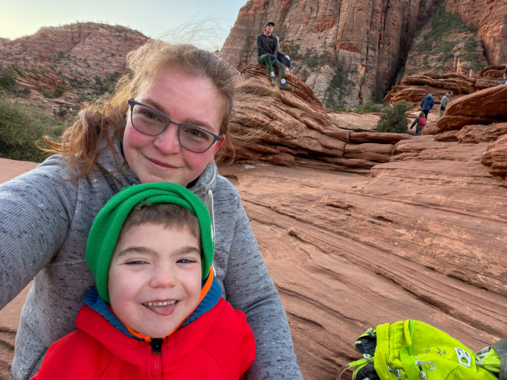 Woman and young child take a selfie on the slickrock trail ascending Angels Landing in Zion National Park during golden morning light.
