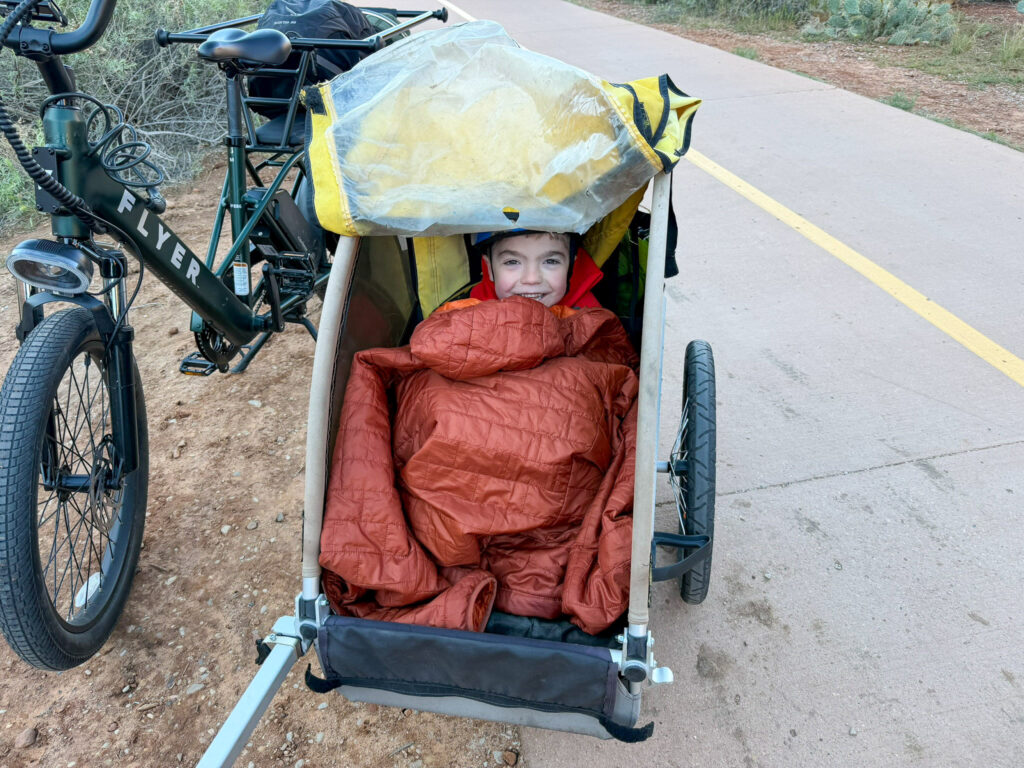 Young child bundled in an orange sleeping bag grins from a bike trailer on Zion National Park's paved Pa'rus Trail.