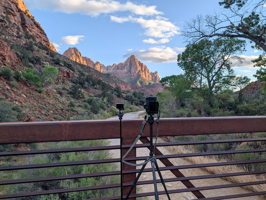 Camera and tripod set up on Watchman Bridge as warm late-day light washes over the Virgin River and red sandstone peaks in Zion National Park.