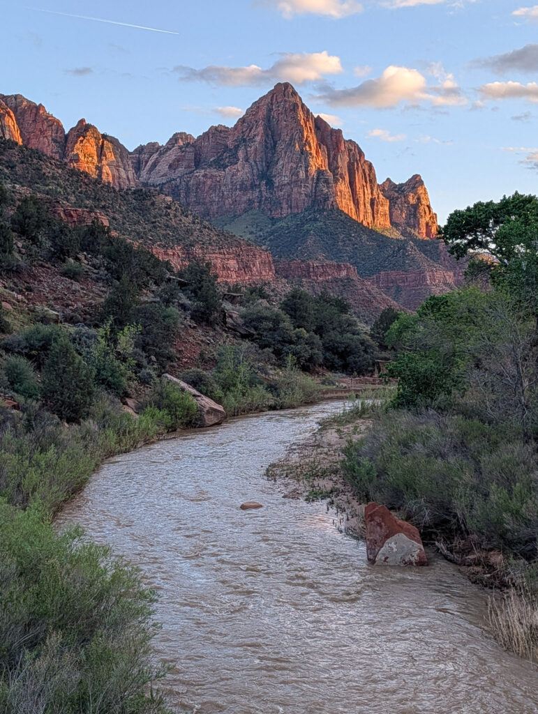 The Watchman glows orange-red in late evening sun above the muddy, flowing Virgin River in Zion National Park.