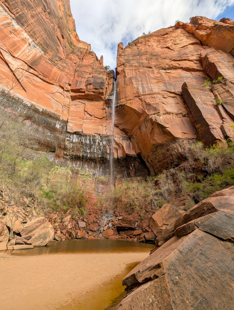 Upper Emerald Pool Falls cascades down towering Navajo sandstone streaked with desert varnish into a turbid reddish-brown pool below.