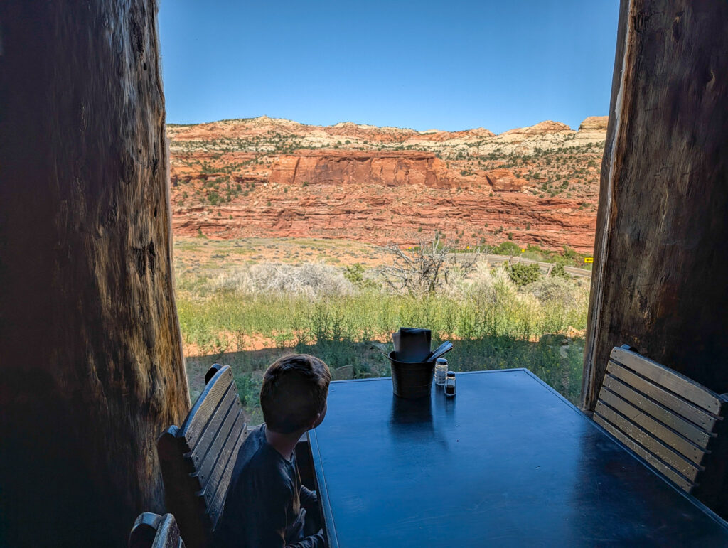 A boy sits at a wooden table inside Kiva Koffeehouse on Utah Scenic Byway 12 looking out an arched window framed by timbers at red sandstone cliffs and grass.
