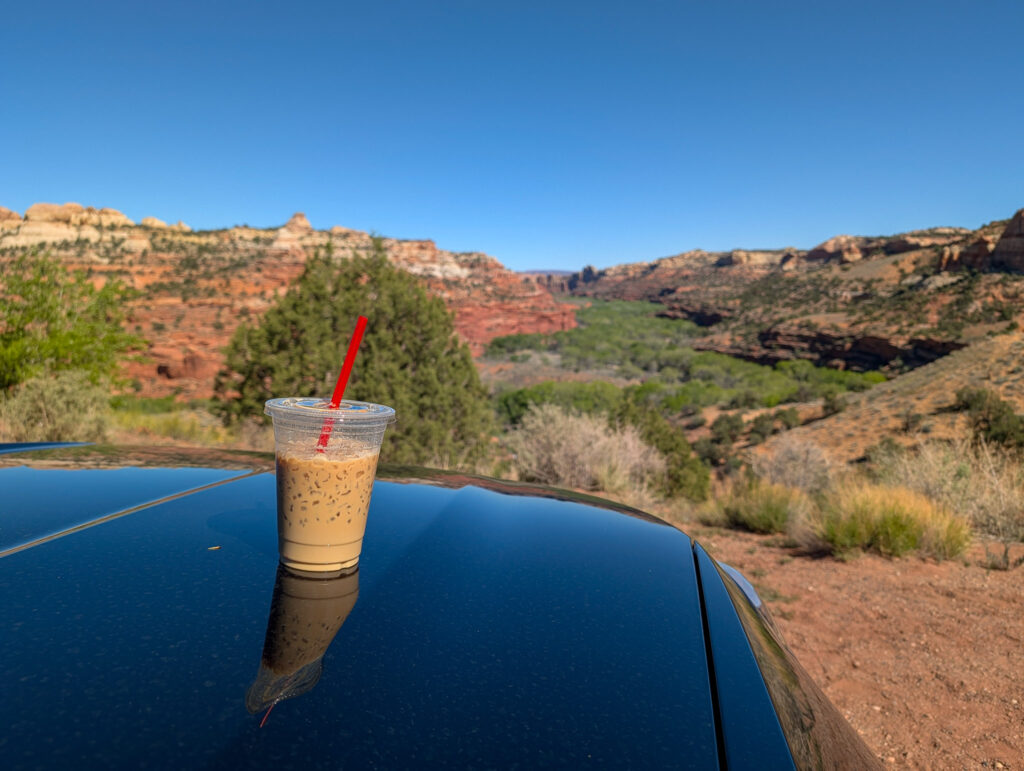 A plastic cup of iced coffee with a red straw rests on the glossy roof of a black car overlooking the red rock canyon below Kiva Koffeehouse on Utah Scenic Byway 12.