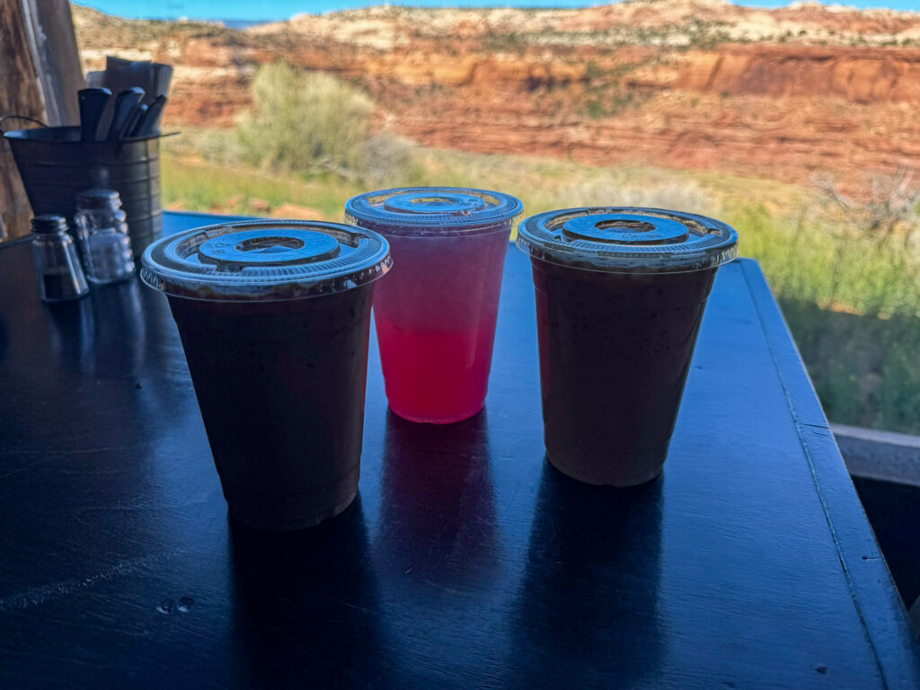 Closer look at three iced drinks on the dark counter at Kiva Koffeehouse on Utah Scenic Byway 12, with the red rock canyon and sage backdrop visible behind.