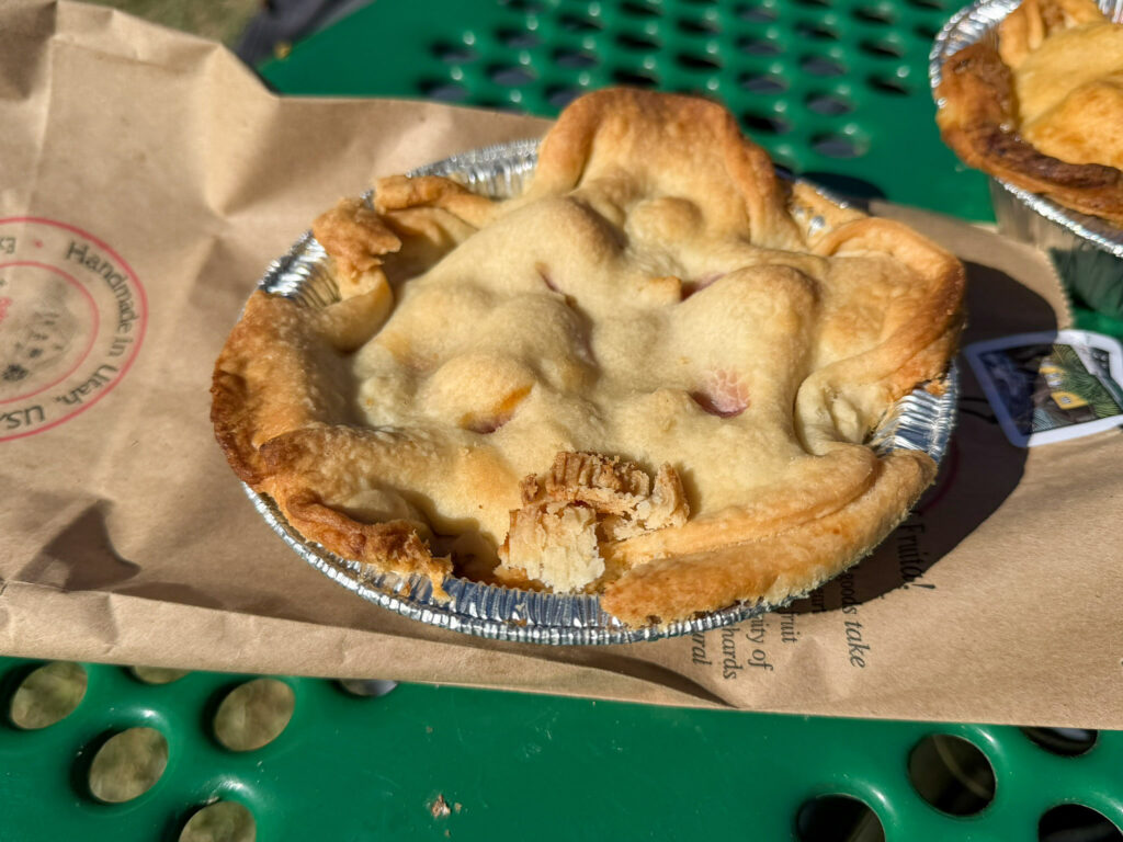A close-up of a freshly baked Gifford House mini peach pie in a foil tin resting on a 'Handmade in Utah' paper bag.