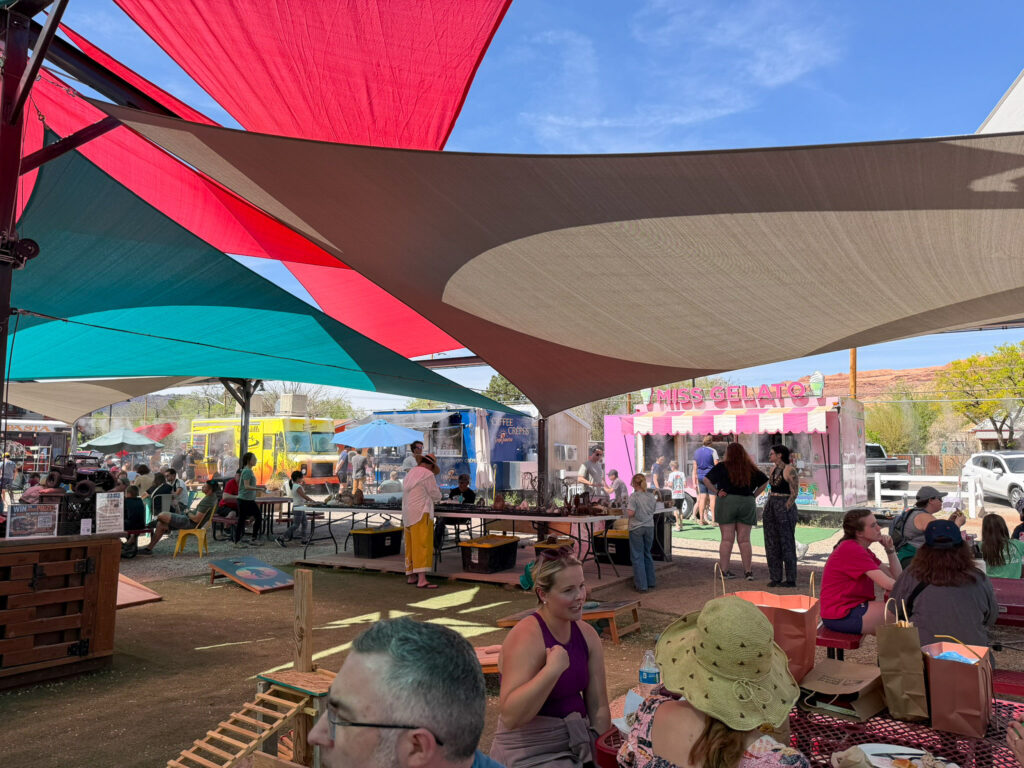 Outdoor food truck plaza in Moab featuring Miss Gelato's pink-striped truck, colorful triangular shade sails, and crowds gathered at picnic tables.