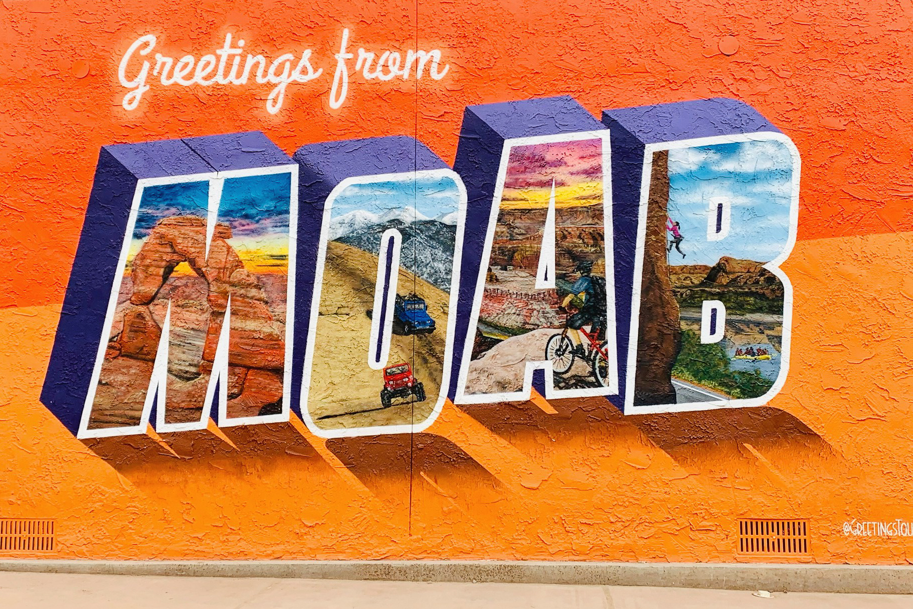 A vibrant orange street mural reading "Greetings from MOAB" in large 3D block letters. Each letter contains a painted scene of Moab's iconic activities and landscapes: Delicate Arch at sunset, off-road jeeping on slickrock, mountain biking on canyon trails, rock climbing, and Colorado River rafting.