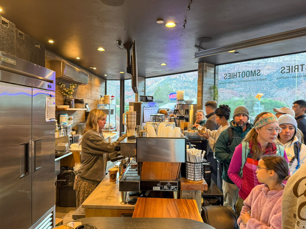 Interior of Canyon Coffee in Springdale, Utah during a busy morning rush with a barista pulling shots at the espresso machine and a long line of customers waiting at the counter.