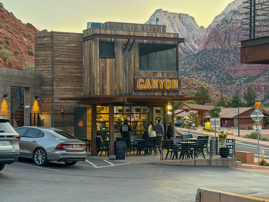 Exterior of Canyon Coffee in Springdale, Utah at dusk with the lit-up wooden 'CANYON' sign over the patio, customers seated outside, and Zion cliffs glowing behind.