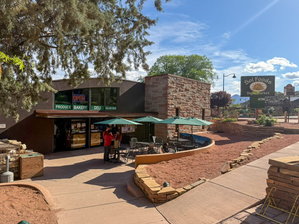 Exterior of Sol Foods Supermarket in Springdale, Utah showing the produce, bakery, and deli entrance with green umbrellas and patio seating in front under a bright sunny sky.