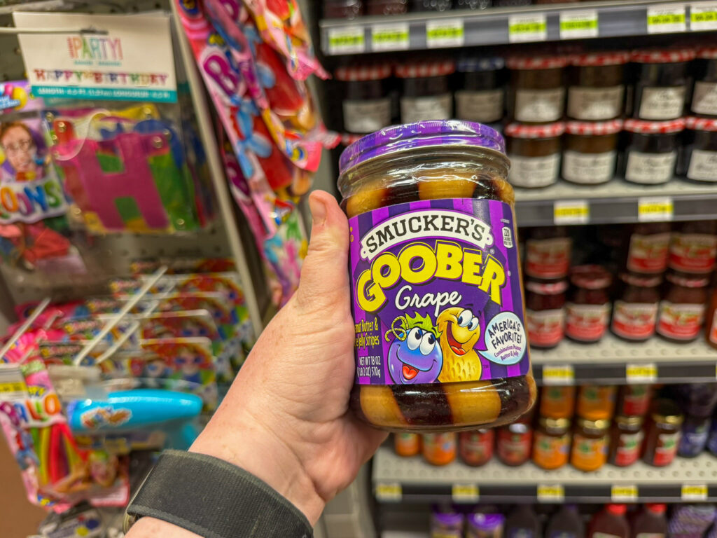 A hand holding a jar of Smucker's Goober Grape peanut butter and grape jelly stripes in the jam aisle at Sol Foods Supermarket in Springdale, Utah, with rows of jars and party supplies behind it.