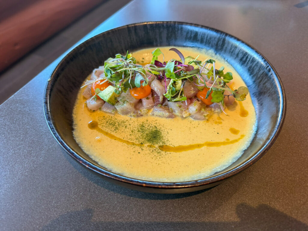 A black ceramic bowl of ceviche at Anu Restaurant in Springdale, Utah with cubed white fish in golden leche de tigre, microgreens, peppers, and a green powder garnish.