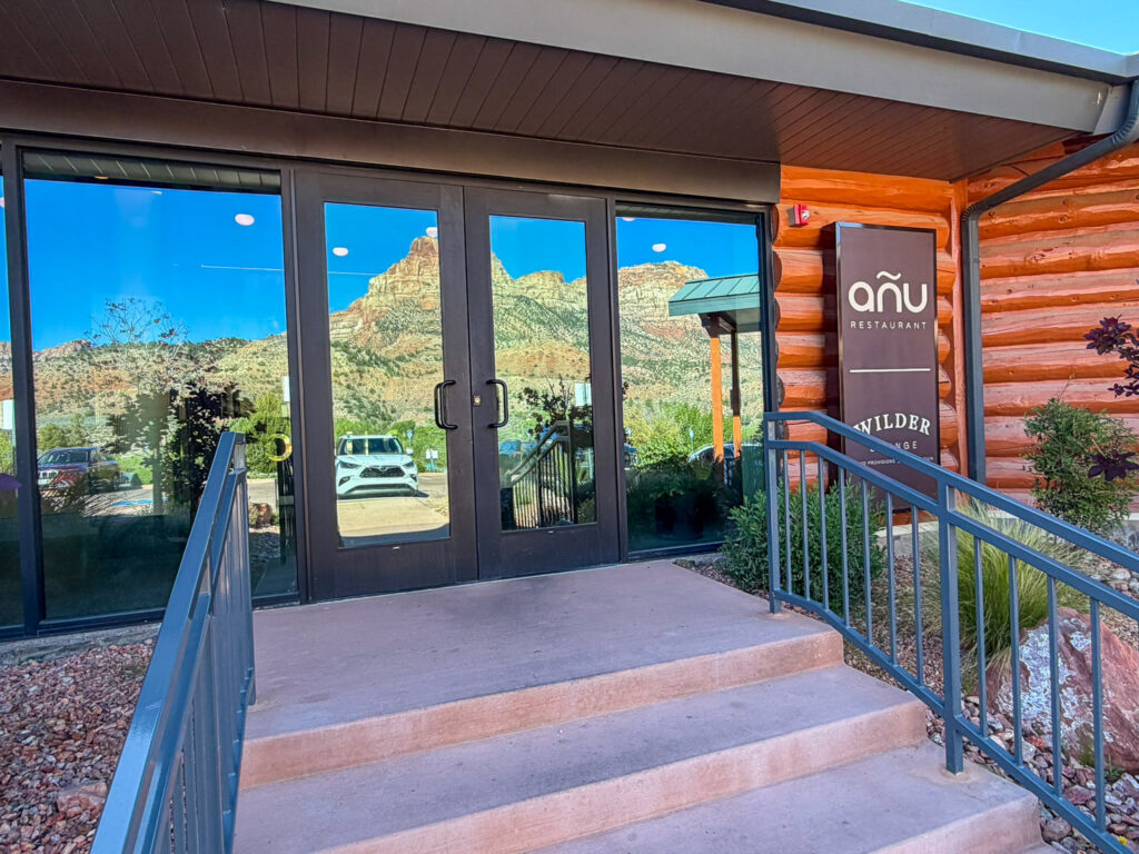 Front entrance steps and glass doors of Anu Restaurant in Springdale, Utah with the wooden Anu signage on the right and the red Zion cliffs reflected in the glass doors and side windows.