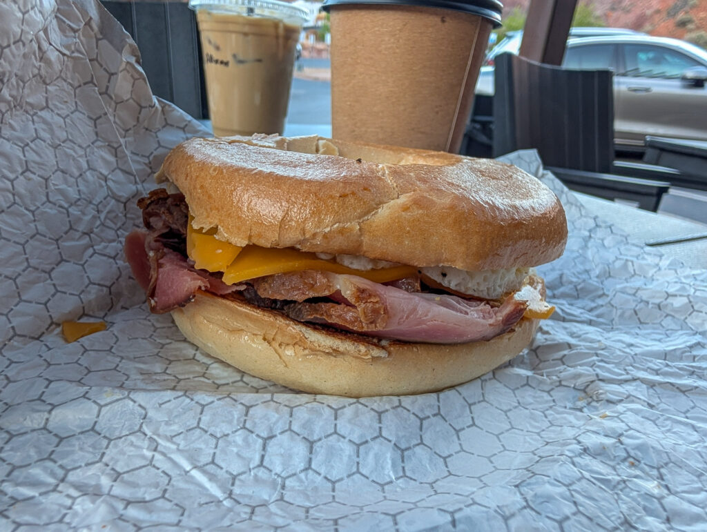 A toasted bagel breakfast sandwich filled with ham, egg, and cheese on parchment paper at Hangry Goat in Springdale, Utah, with iced and hot coffees behind it on the patio.
