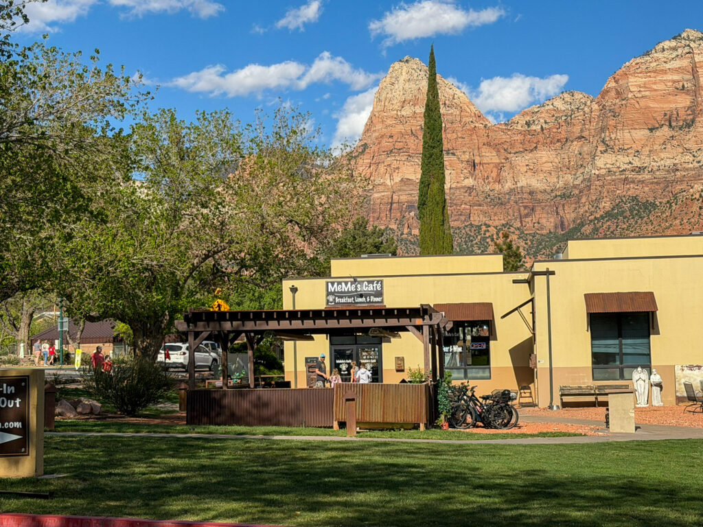 MeMe's Cafe in Springdale, Utah viewed from the lawn with the towering red sandstone cliffs of Zion National Park rising behind the cafe under a partly cloudy sky.