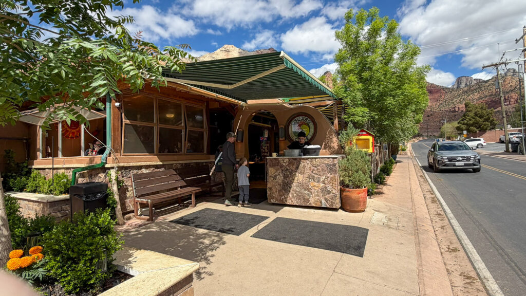 Sidewalk view of Oscar's Cafe storefront in Springdale, Utah with the green awning, wooden bench, and Zion cliffs rising behind the building under a bright partly cloudy sky.