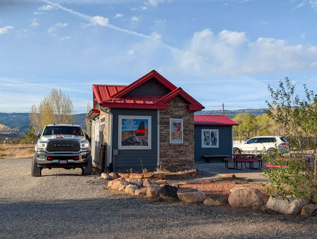 Morning view of the small red-roofed Dark Sky Coffee shop in Torrey, Utah with a pickup truck parked at the drive-through window and red cliffs in the distance.