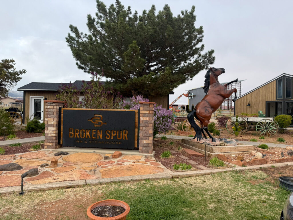 The Broken Spur Inn entrance sign with a bronze rearing horse statue, pine tree, and garden under an overcast sky in Torrey, Utah.