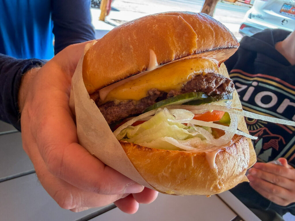 Close-up of a thick cheeseburger with melted American cheese, lettuce, tomato, and onion on a brioche bun, held in a paper wrapper at Capitol Burger food truck in Torrey, Utah.