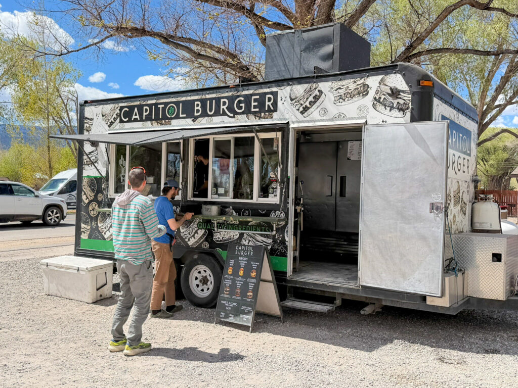 The Capitol Burger food truck in Torrey, Utah with two customers ordering at the window beside a chalkboard menu, surrounded by trees against a blue sky.