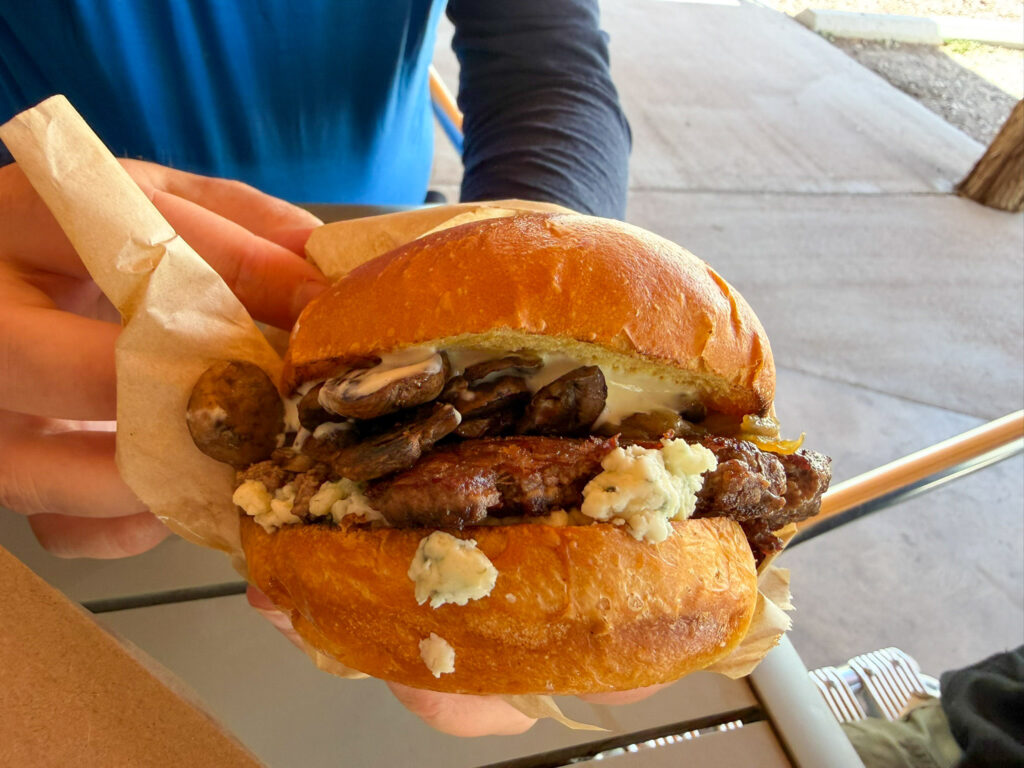A thick burger topped with sautÃ©ed mushrooms, crumbled blue cheese, and a creamy sauce on a brioche bun, held in paper wrapping at Capitol Burger food truck in Torrey, Utah.
