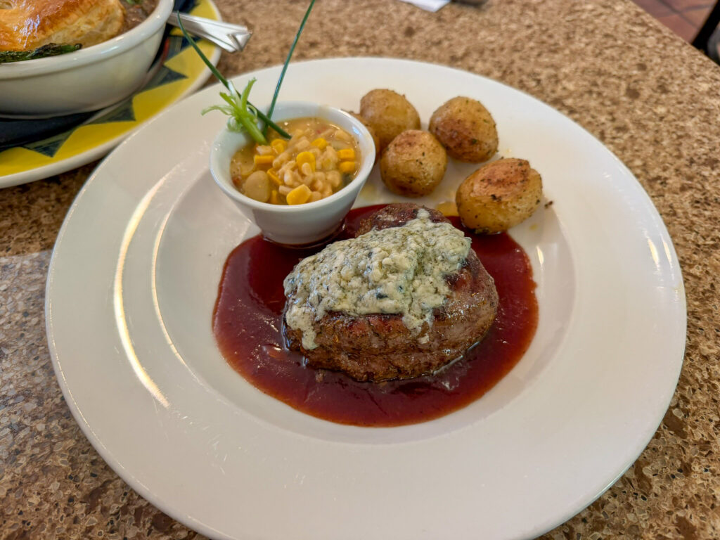 A filet steak topped with melted blue cheese in red wine reduction, served with crispy potato puffs and a small bowl of buttery corn on a wide white plate at Hunt and Gather restaurant in Torrey, Utah.