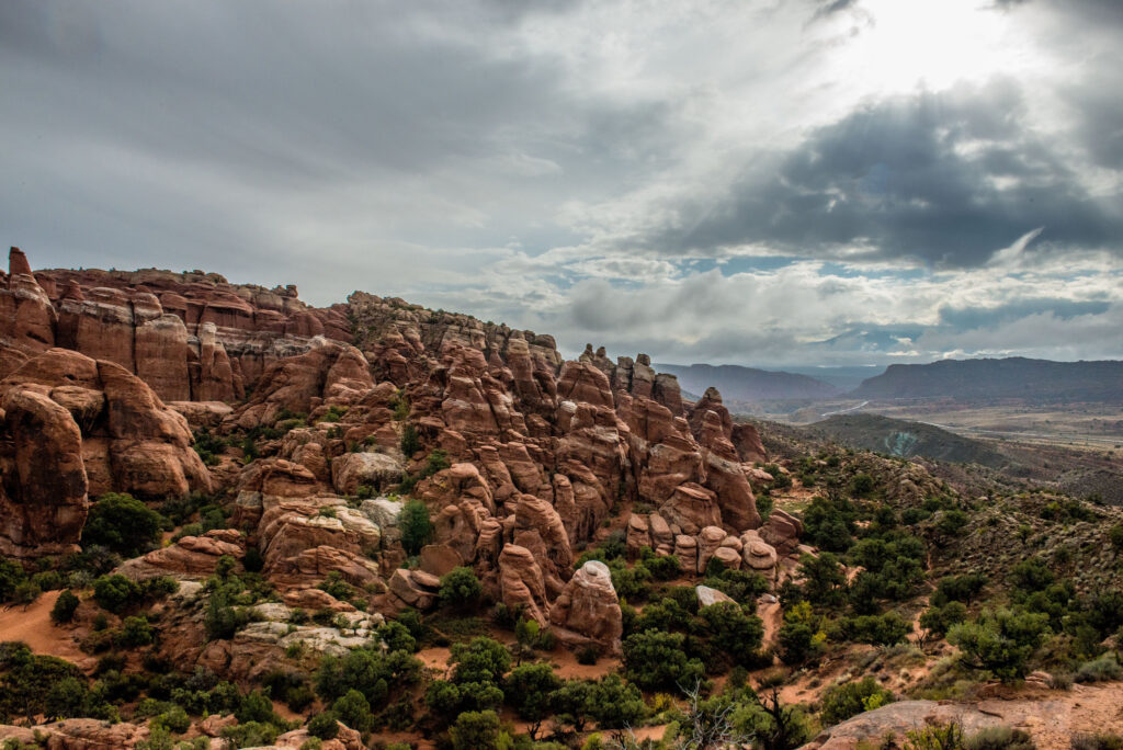 Densely packed red sandstone fins and rounded formations of Fiery Furnace rise above a shrub-dotted slope, with a broad valley and distant mesas beneath a dramatic overcast sky.
