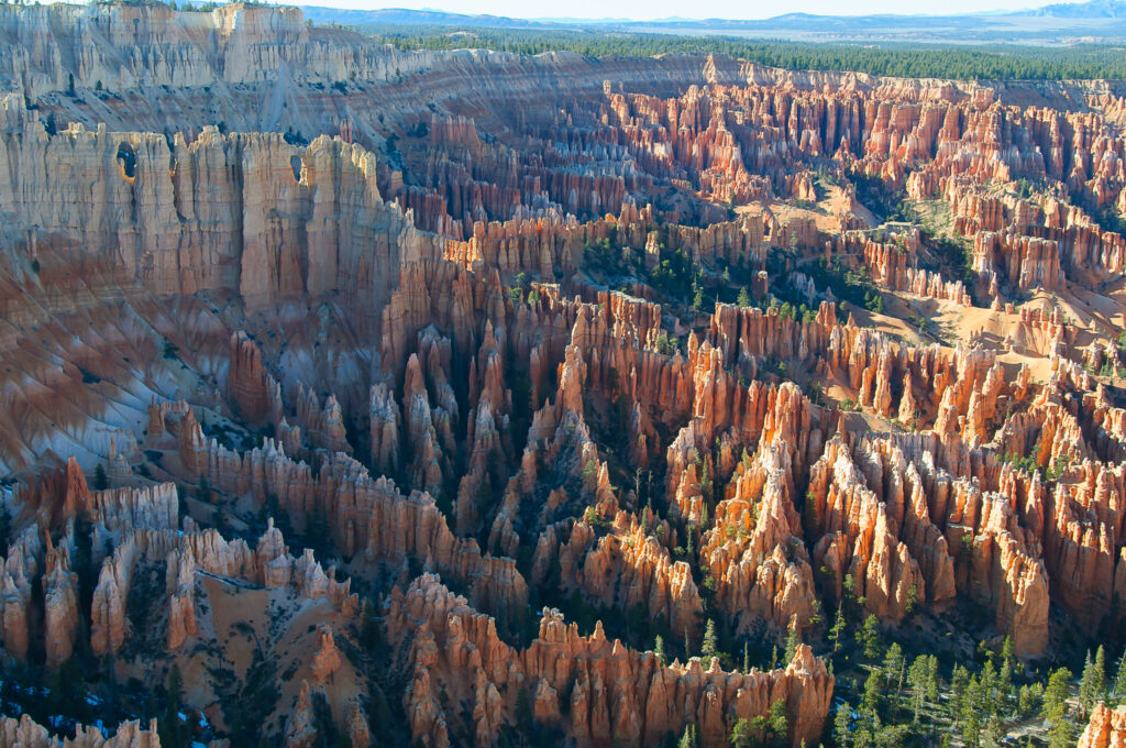 Aerial view from Bryce Point looking down into Bryce Amphitheater, where dense clusters of orange, red, and white hoodoos fill the canyon, interspersed with conifers, beneath a pale blue sky.