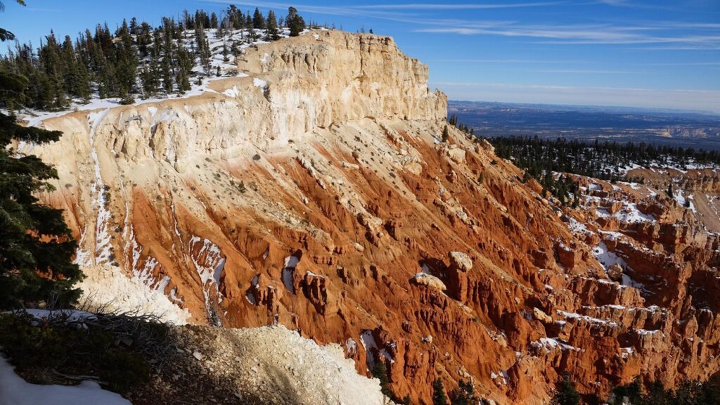 Bristlecone Loop trail at Bryce Canyon, with a cream-and-orange banded sandstone butte, snow-dusted slopes, red hoodoos receding into the canyon, and a distant mesa under blue sky.