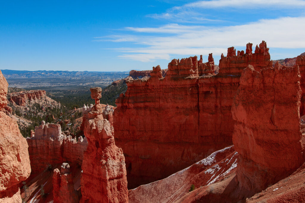 Sweeping view of Bryce Canyon's Navajo Loop, with Thor's hammer and a dramatic cliff wall rising above snow-dusted ledges, conifers on the canyon floor, and distant mountain ranges under a bright blue sky.