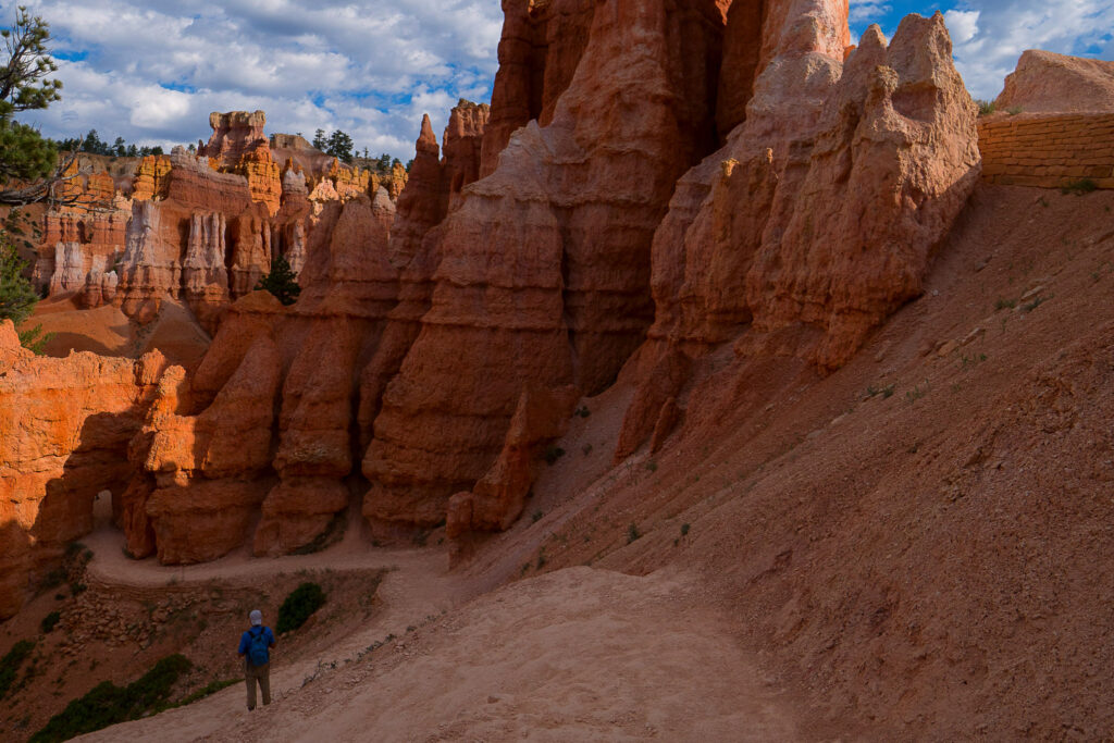 A hiker in a blue jacket gazes up at towering orange and red hoodoos lining the Queen's Garden trail in Bryce Canyon, with pine trees and a partly cloudy sky above.