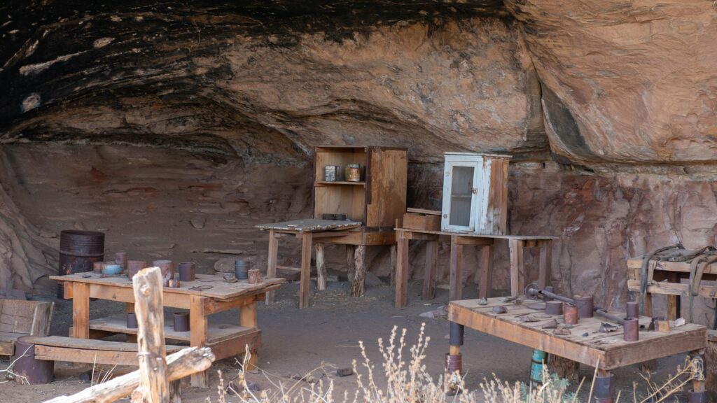 A sandstone alcove at Cave Spring in Canyonlands shelters a preserved cowboy camp with rustic worktables, tin cans, tools, wooden cabinets, and a rusted barrel beneath a soot-darkened rock ceiling.